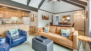 A spacious living area with a leather sofa, a blue armchair, and a coffee table. There is a kitchen in the background with wooden cabinets and a refrigerator.