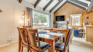 A cozy, well-lit dining area with wooden furniture, a television above the fireplace, and a large window offering a view of the outdoors.