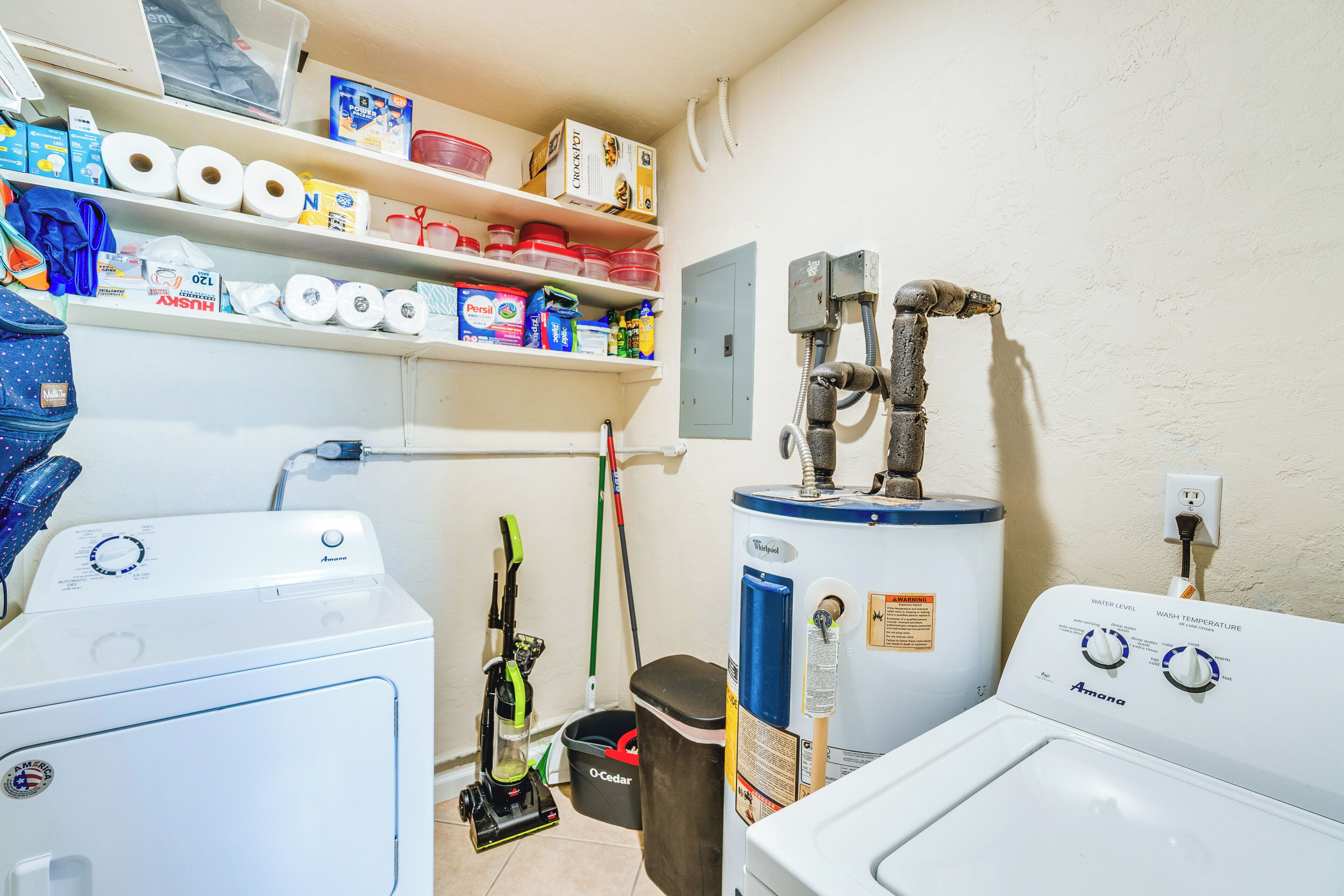 A laundry room with two washing machines, a water heater, and shelves stocked with various supplies.