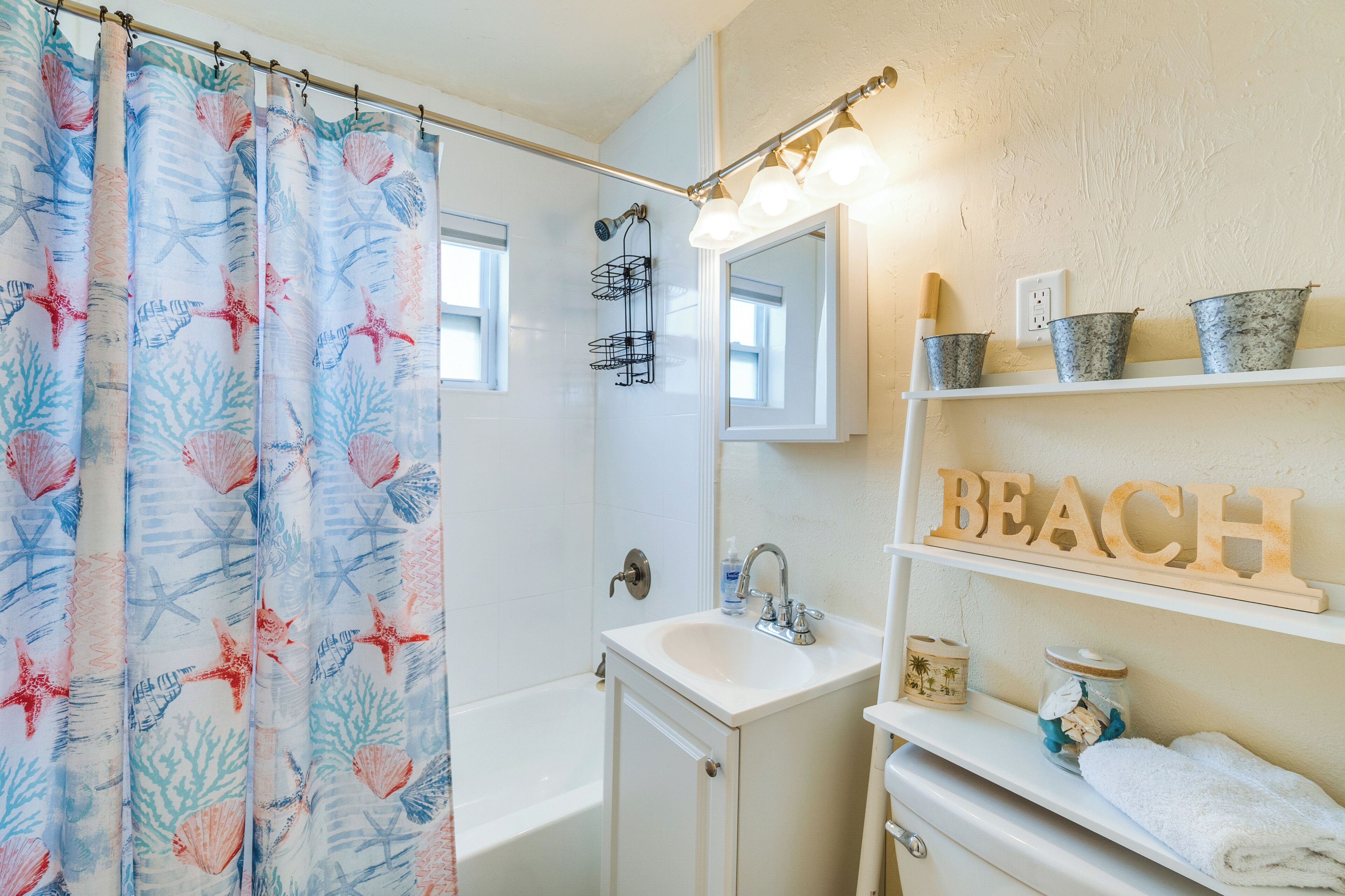 Un baño con cortina de ducha con estampado de vida marina, una bañera, un lavamanos con un espejo encima y estantes con objetos decorativos, incluyendo un letrero que dice 'BEACH'.