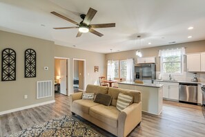 Ein modernes Wohnzimmer mit einem beige farbenen Sofa, einem Deckenventilator und einer Küchenzeile mit weißen Schränken und Edelstahlgeräten.