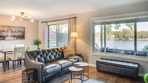 A modern living room with a leather sofa, a dining table with chairs, a lamp, and a large window with a view of a body of water. - Riverfront Champlin Townhome w/ Deck & Water View! (Champlin)