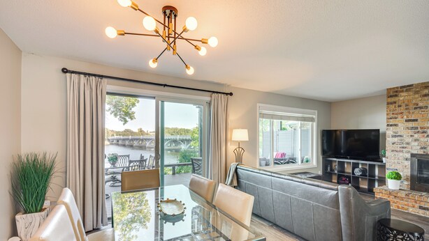 A modern living room with a glass dining table, leather sofa, and a TV mounted on a brick wall. - Riverfront Champlin Townhome w/ Deck & Water View! (Champlin)