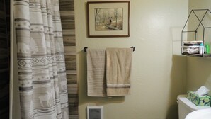 Une salle de bain avec un rideau de douche, un porte-serviettes avec deux serviettes, un tableau encadré au mur, une grille d’aération et une étagère avec des articles de toilette.