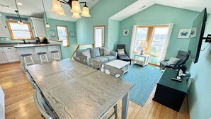 A spacious living area with a large wooden dining table, a gray sofa, and a black armchair. There is a kitchen area with a bar counter and stools, and a large window providing natural light. - KD300- Walk to Beach, Heated Saltwater Pool, Hot Tub (Kill Devil Hills)