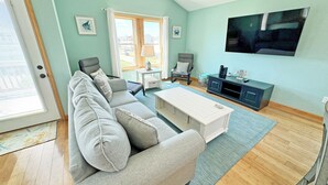 A living room with a grey sofa, a white coffee table, a TV on the wall, and a window with curtains. - KD300- Walk to Beach, Heated Saltwater Pool, Hot Tub (Kill Devil Hills)