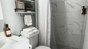 A bathroom with a toilet, a sink, a shower with a glass door, and a shelf with toiletries. - The perfect blend of comfort and convenience in our cozy studio apartment (CAPE CORAL FL)