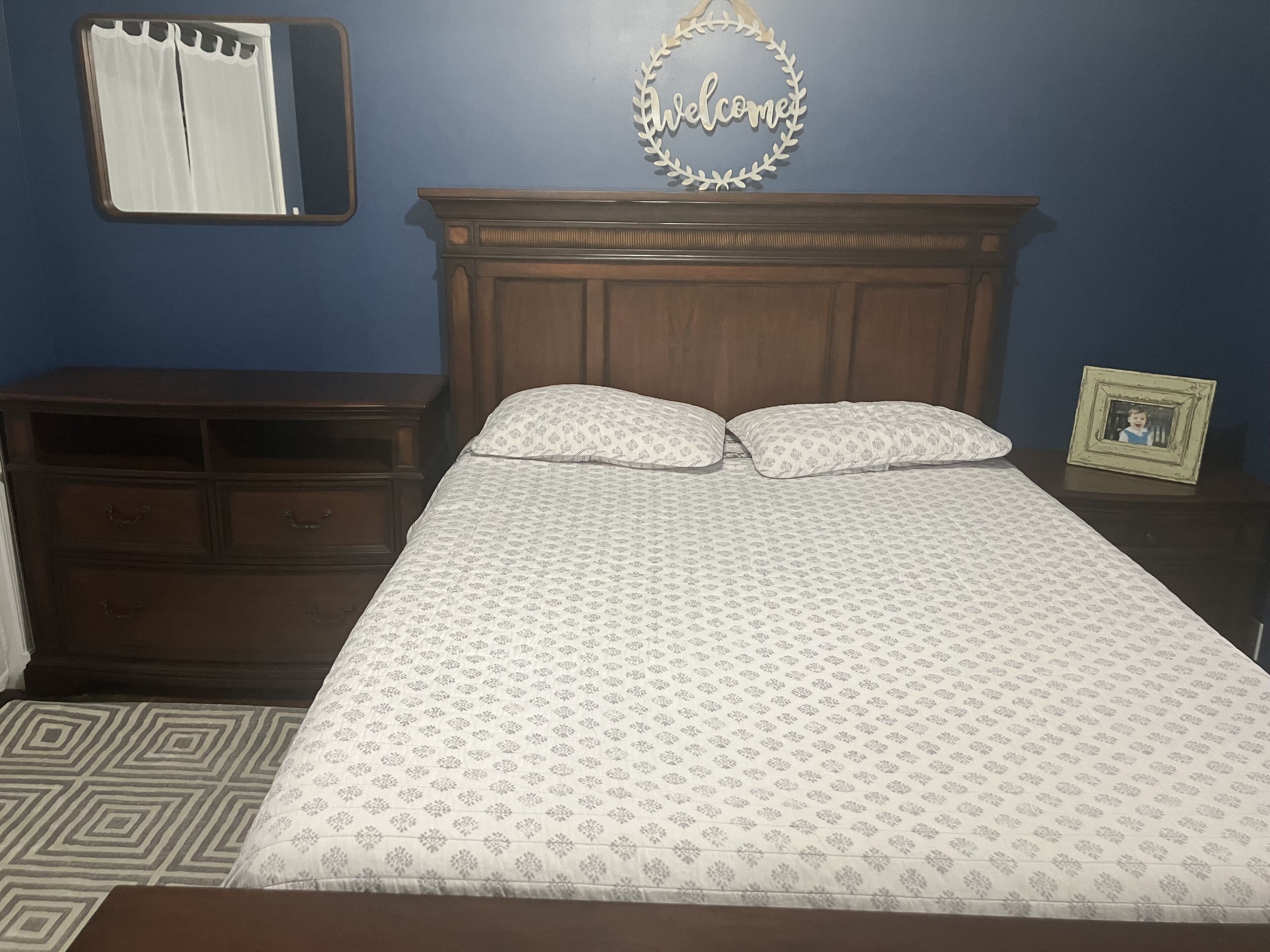A bedroom with a wooden headboard, two bedside tables, a bed with patterned bedding, a mirror, and a 'Welcome' sign on the wall.