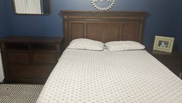 A bedroom with a wooden headboard, two bedside tables, a bed with patterned bedding, a mirror, and a 'Welcome' sign on the wall.