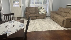 A living room with two sofas, a dining table with a floral tablecloth, and a large window with curtains.