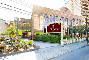 Experience the grand entrance of Corinto Hotel, featuring a striking facade with arches and a balcony area overlooking a beautifully landscaped garden with a serene fountain.