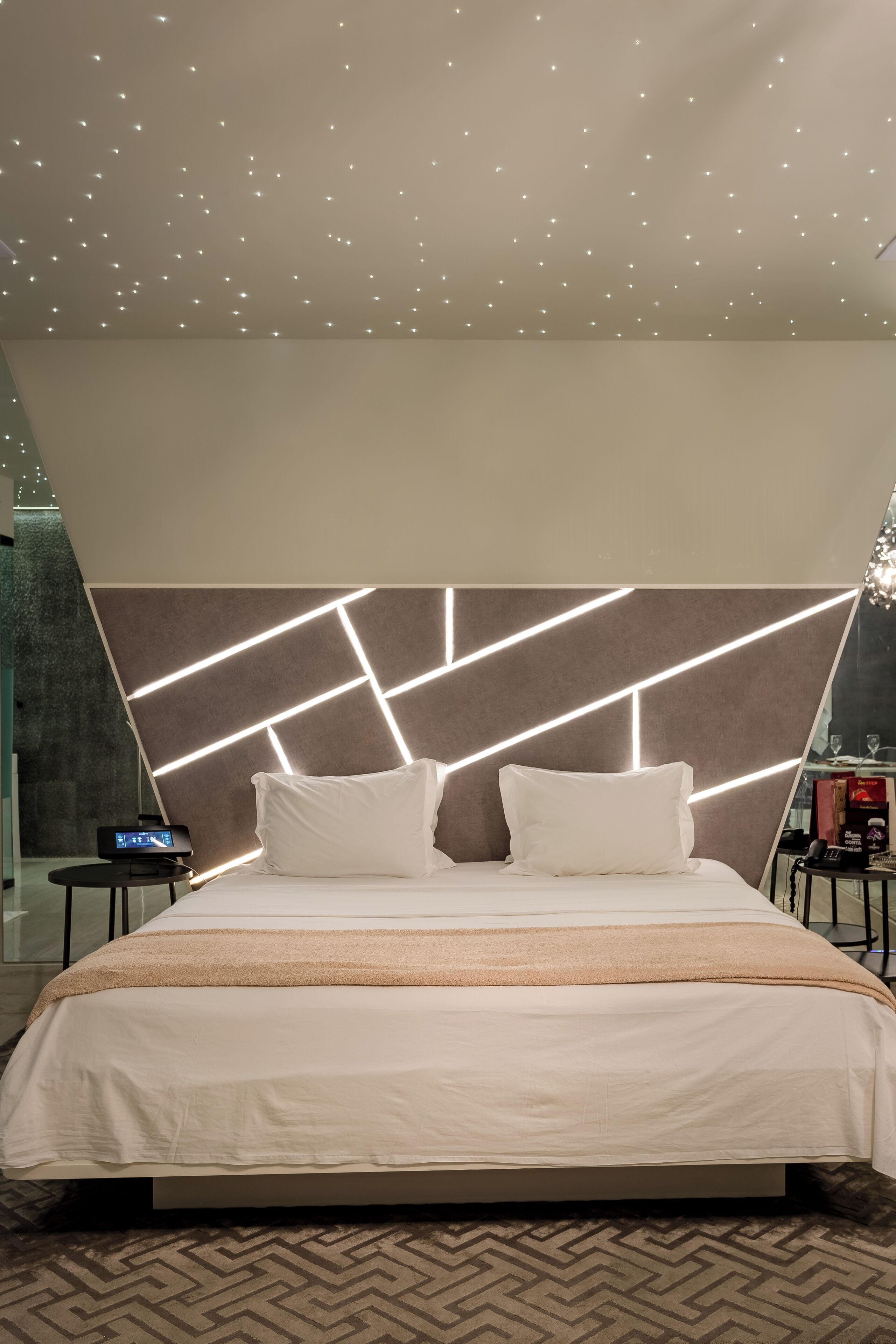 Ein modernes Hotelzimmer mit einem großen Bett, einer Kopfteilbeleuchtung mit geometrischem Design, einem Nachttisch und einem kleinen Beistelltisch.