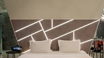 Ein modernes Hotelzimmer mit einem großen Bett, einer Kopfteilbeleuchtung mit geometrischem Design, einem Nachttisch und einem kleinen Beistelltisch.