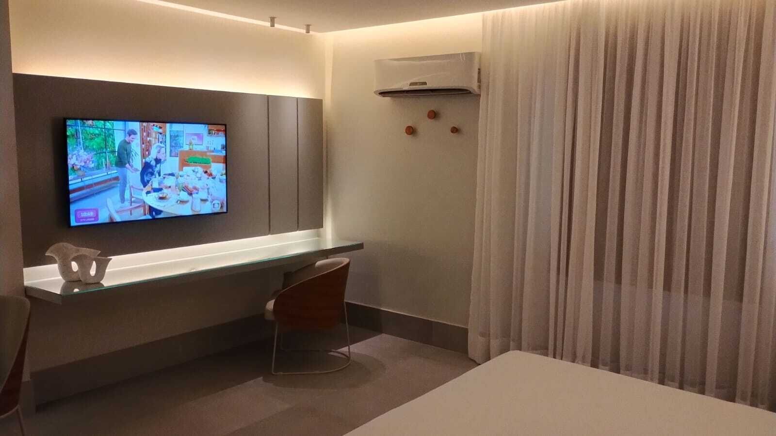 Et hotelværelse med fjernsyn monteret på væggen, et glasbord, en stol, en seng og et vindue med gardiner.