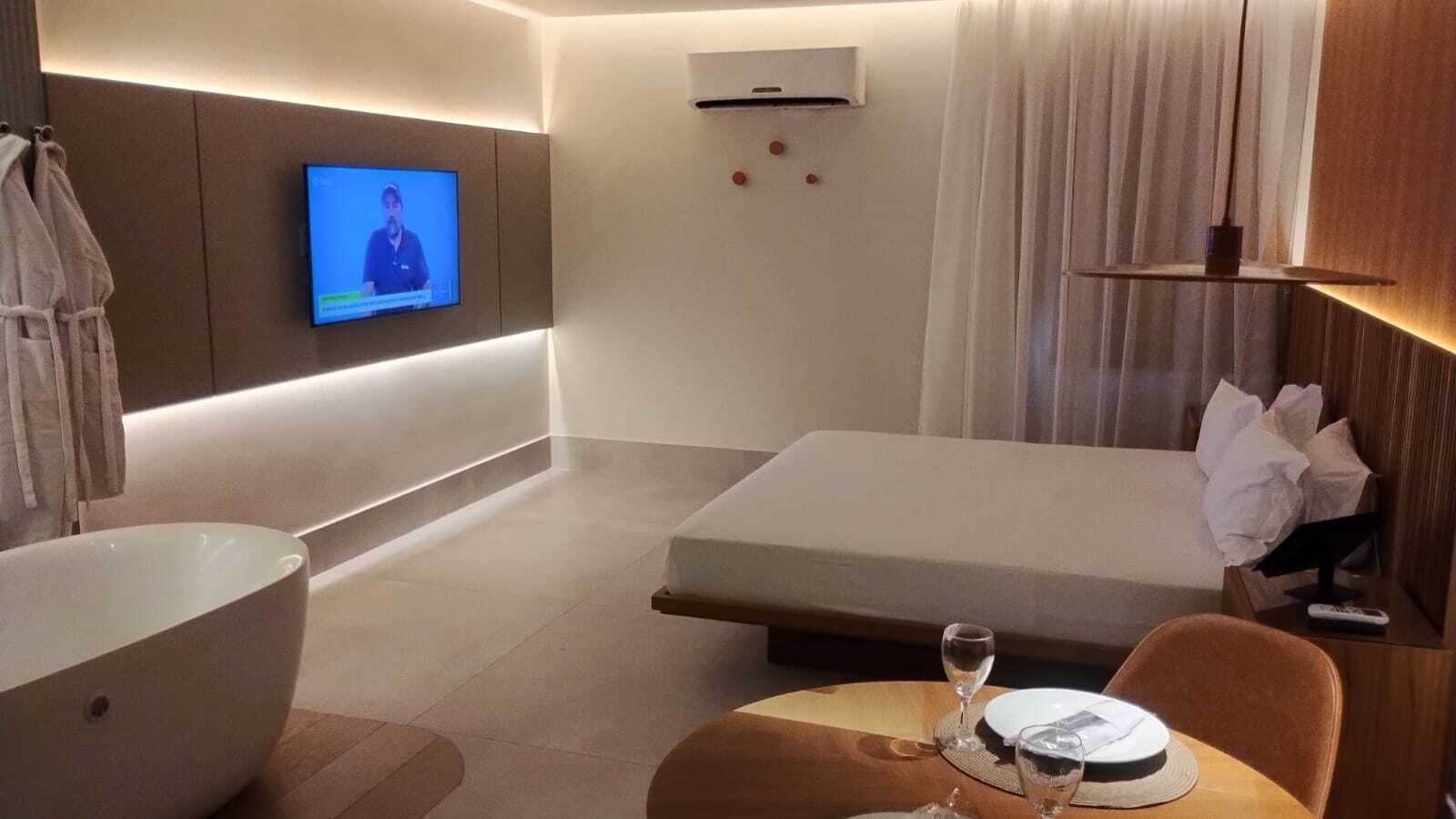 A modern hotel room with a large bathtub, a bed, a TV mounted on the wall, and a small table with a wine glass.