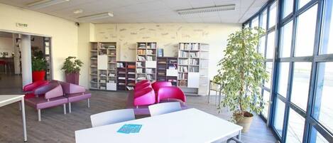 A modern library with white walls, large windows, and bookshelves filled with books. There are colorful chairs and a table in the center.