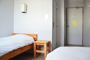 A room with two single beds, a wooden nightstand, a wall-mounted light, and a door with a notice. - La Digue (Calais)