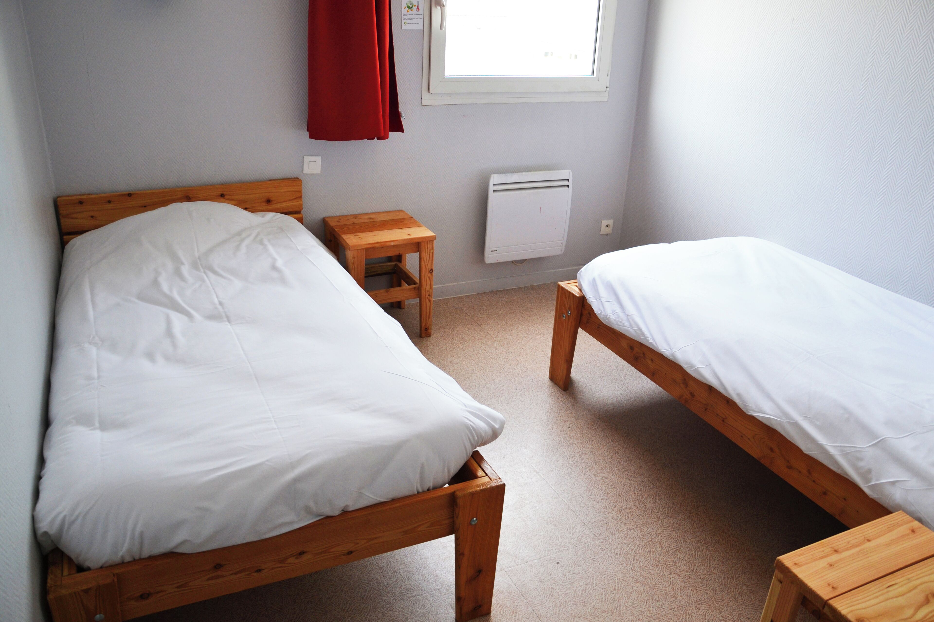 Ein Zimmer mit zwei hölzernen Betten, einem kleinen Holztisch, einem roten Vorhang und einem Fenster mit Tageslicht.