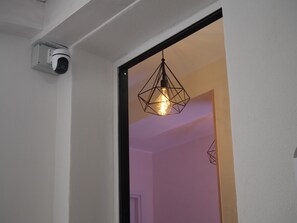 A security camera mounted on a white wall, a geometric pendant light, and a doorway leading to another room.