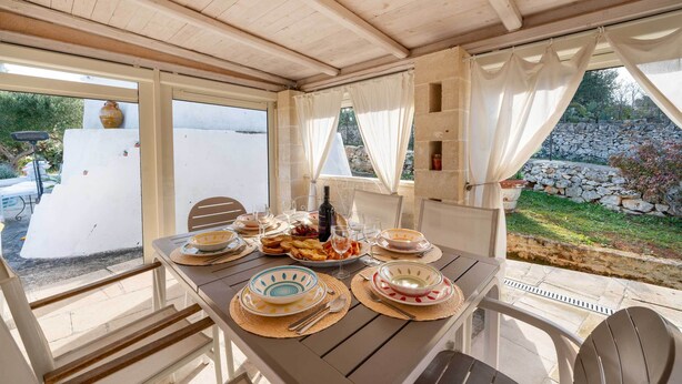 Un área de comedor al aire libre con una mesa lista para una comida, rodeada de sillas y vistas a un jardín y un muro de piedra.