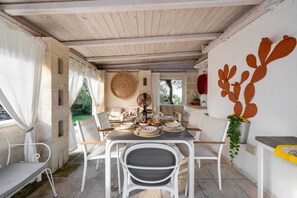 A covered outdoor dining area with a table set for a meal, surrounded by white chairs, and a decorative wall art piece. - 3723 Trullo Bello Sguardo by Barbarhouse (Ostuni)