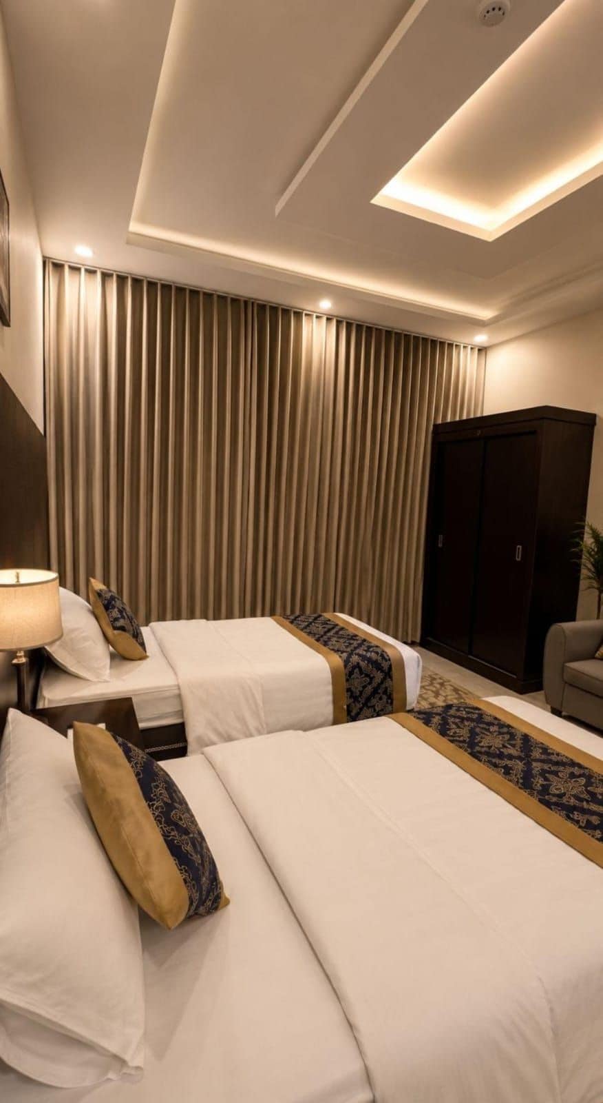 Quarto de hotel com duas camas, um guarda-roupa, um sofá e um abajur.