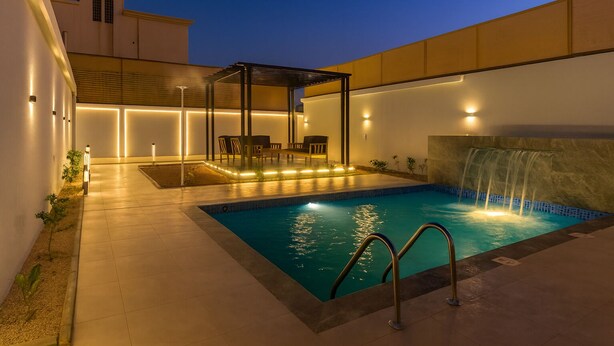 A swimming pool with a waterfall feature, surrounded by a covered seating area with chairs and tables, and illuminated by ambient lighting. - تميُّز الاقامة للشقق المخدومة Tamyoz Aqama for Serviced Apartments (Ha'il)