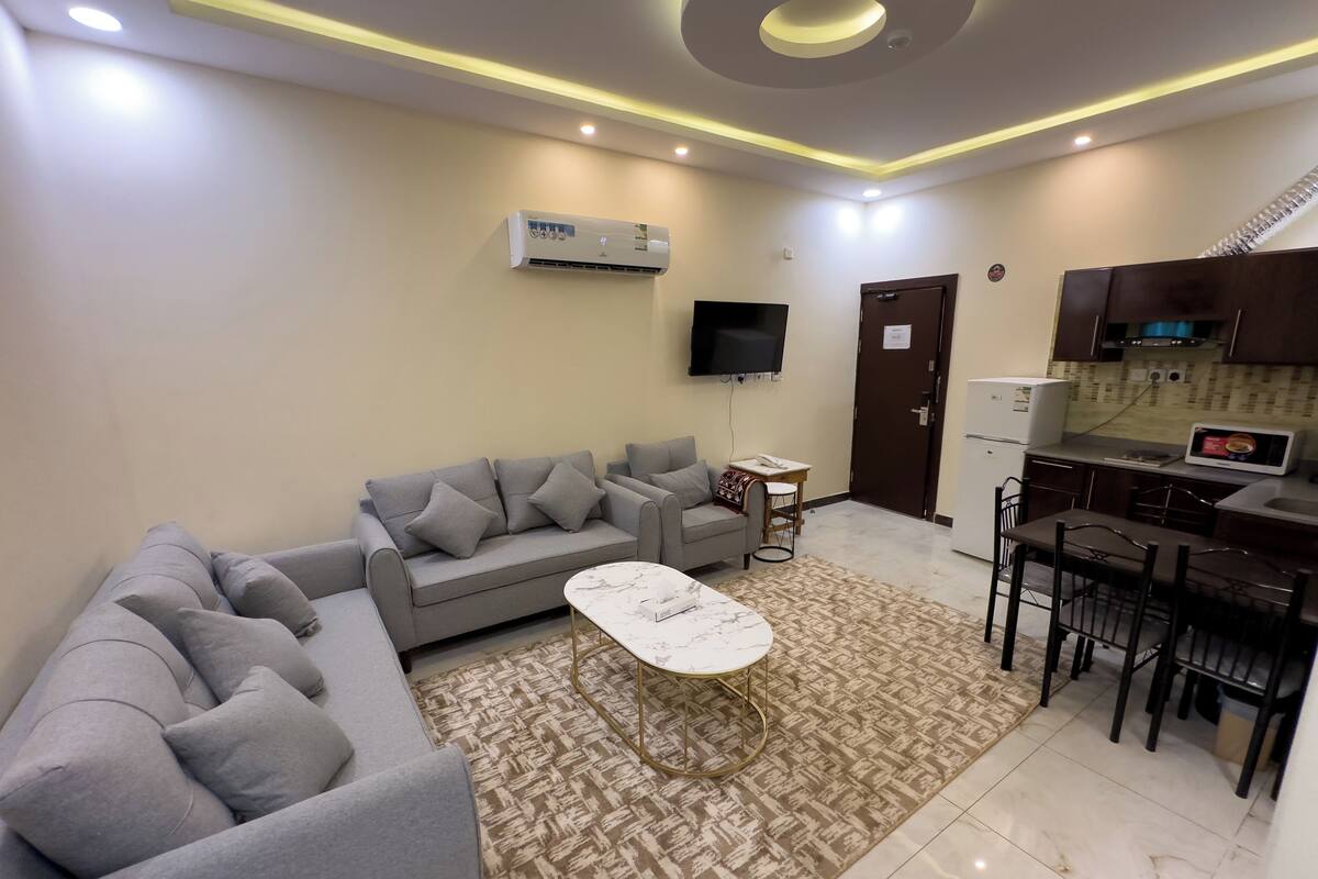 Uma sala de estar moderna com um sofá seccional cinza, uma mesa de centro branca e uma área de cozinha com micro-ondas e fogão.