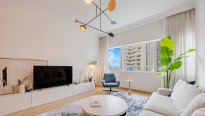 Una sala de estar moderna con una televisión de pantalla plana, una mesa de centro redonda, un sillón azul, un sofá blanco y un ventanal con vistas a la ciudad.