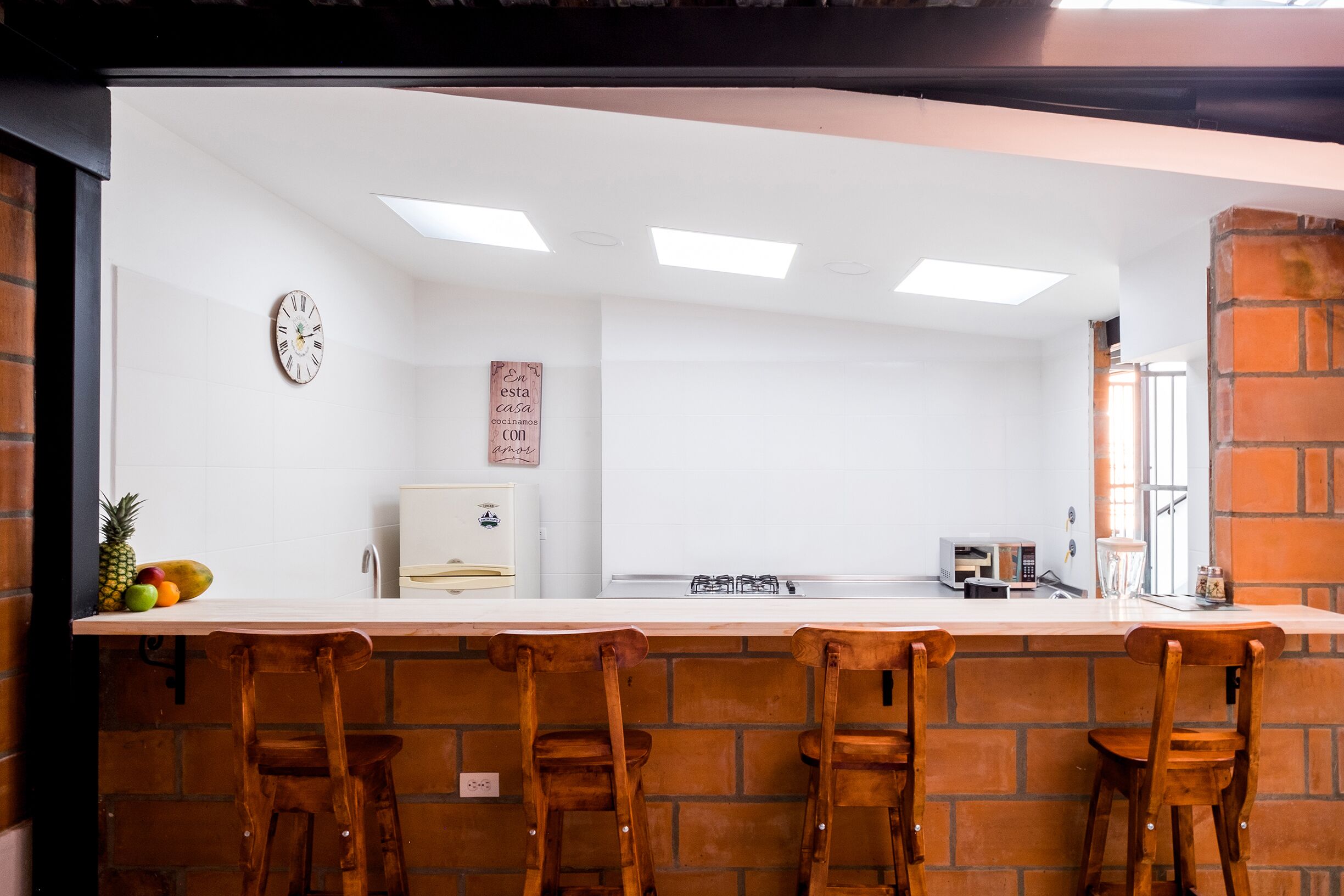 Una cocina con encimera blanca, cuatro taburetes de madera, un reloj y un refrigerador.