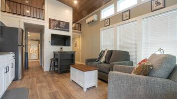 Una sala de estar moderna con un sofá gris, una mesa de centro blanca y un televisor montado en la pared. Se ve una escalera que conduce a un nivel superior, y un área de cocina al fondo.