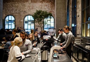 Un café animé, avec des clients assis à des tables, un barista préparant des boissons et une grande machine à café.
