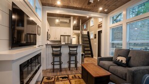 Una cocina moderna con armarios blancos, una isla central con taburetes, una chimenea y un comedor con una escalera que sube a un nivel superior.