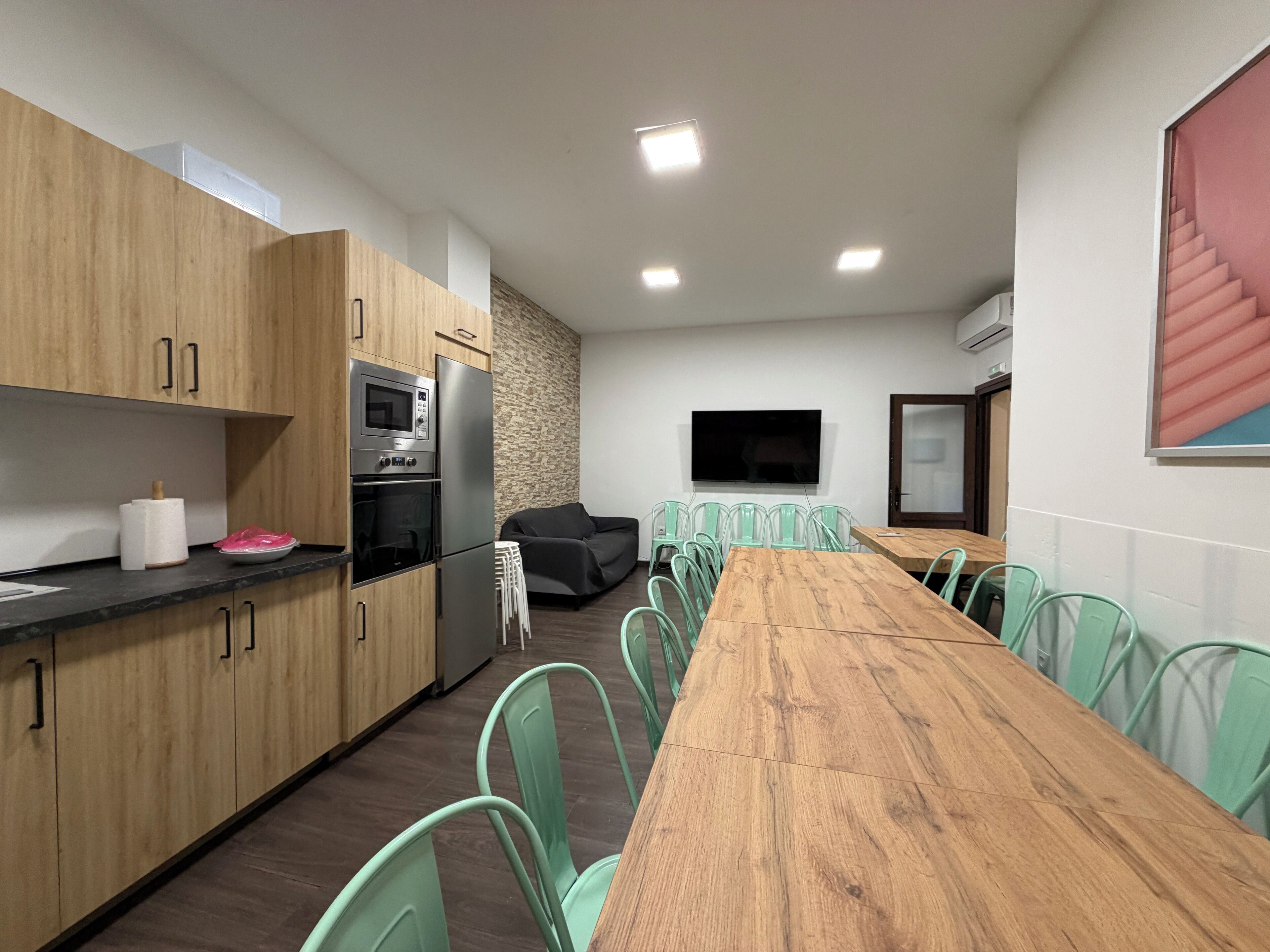 Une cuisine moderne avec des armoires en bois, un four encastré et un four à micro-ondes. On y trouve un coin repas avec une longue table en bois et des chaises vertes. Un téléviseur à écran plat est fixé au mur, et un escalier est visible au fond.