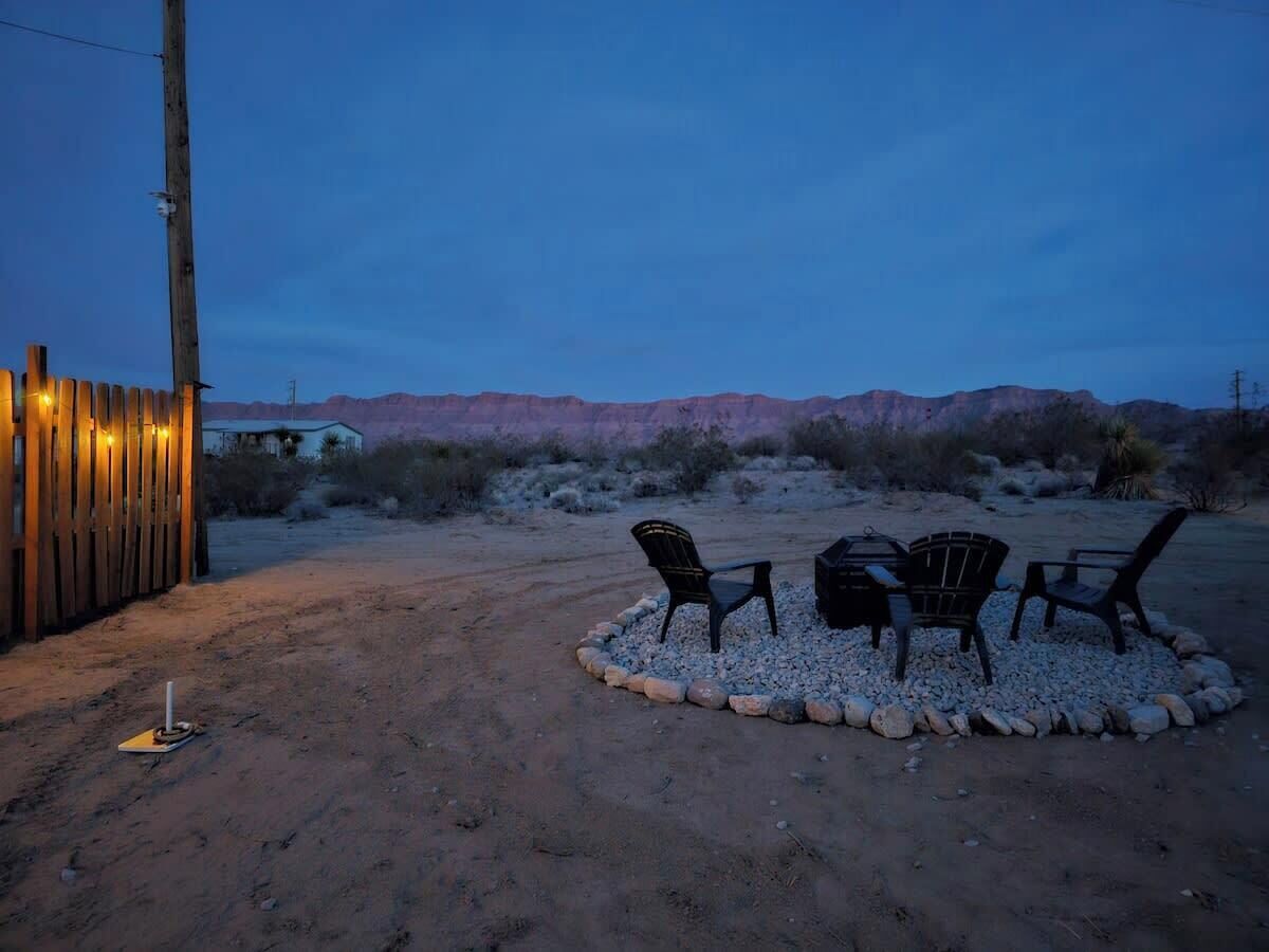 An outdoor seating area with chairs and a table, surrounded by a gravel circle, against a backdrop of desert hills and a wooden fence.