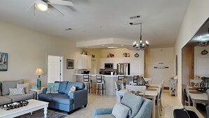 A spacious living room with a sofa, armchair, and coffee table. A kitchen with white cabinets and a dining area with a table and chairs are visible in the background. - Pavilion Palms #201A | Central Location + Close to Beach! (Miramar Beach)