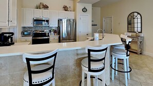 A modern kitchen with white cabinets, stainless steel appliances, and a bar area with stools. - Pavilion Palms #201A | Central Location + Close to Beach! (Miramar Beach)