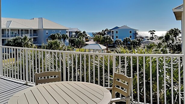 A balcony with a table and chairs, overlooking a beachfront with buildings and palm trees. - Pavilion Palms #201A | Central Location + Close to Beach! (Miramar Beach)