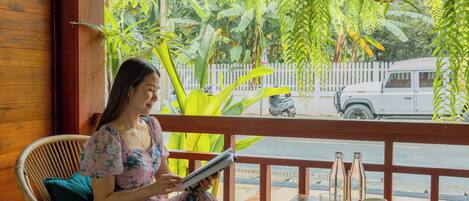 A woman sitting on a wicker chair reading a book on a porch with a wooden table, a white fence, and greenery in the background.