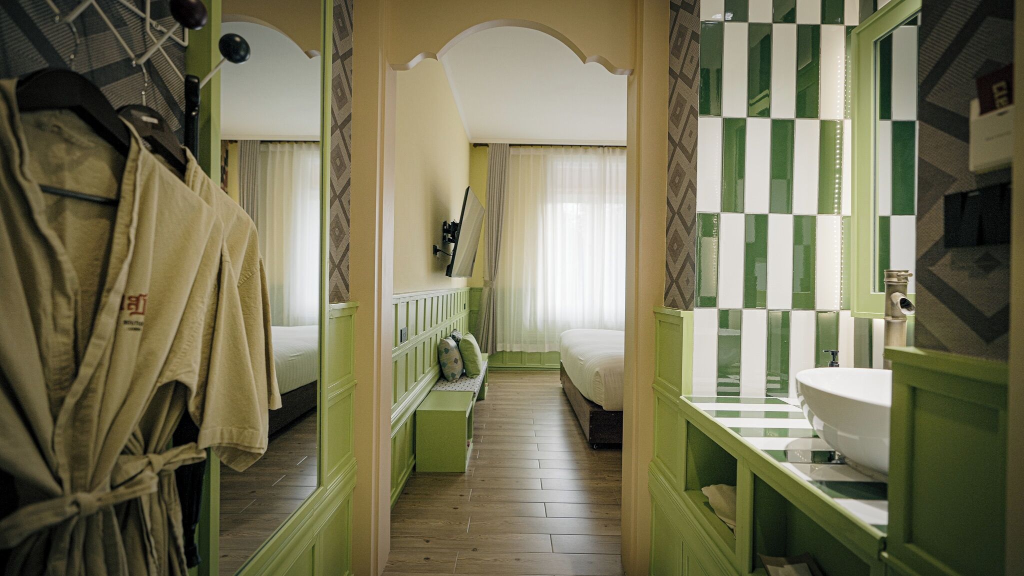 Un pasillo estrecho con una cama, un baño con salpicadero de azulejos blancos y verdes a cuadros, y un perchero.