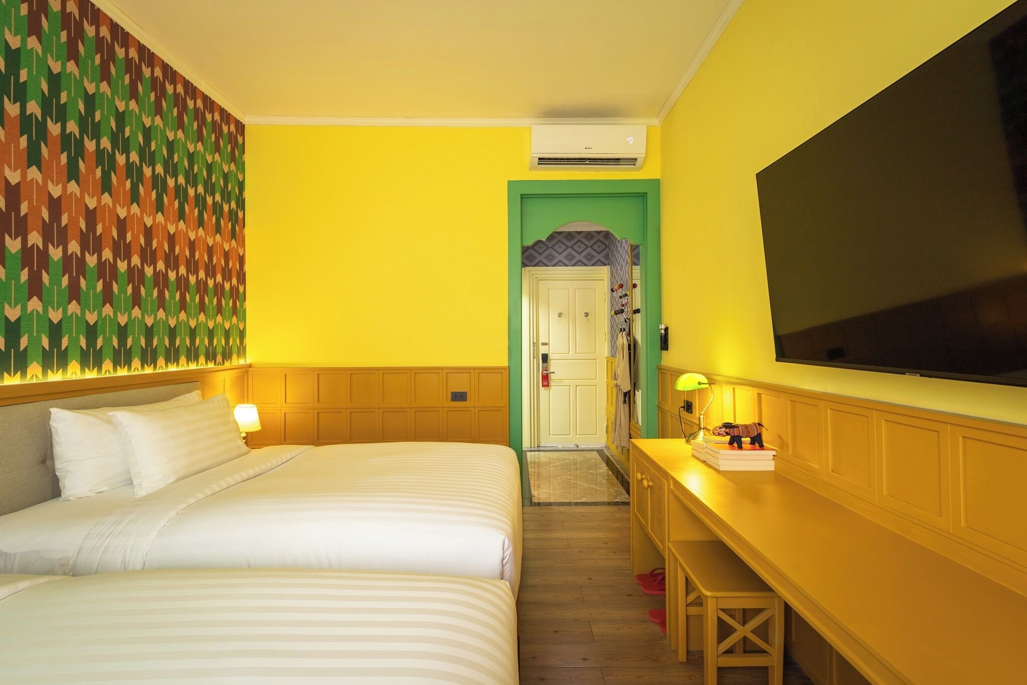 A hotel room with two beds, a television, a desk with a lamp, and a patterned wall.