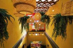 Un escalier orné de lampes suspendues en osier et de fougères retombantes, menant à une pièce dotée d’une fenêtre rouge et d’une fresque murale aux couleurs vives.