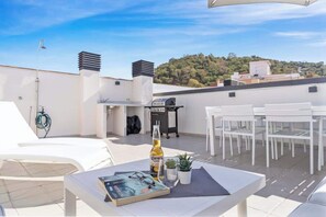 Eine Dachterrasse mit weißen Gartenmöbeln, einem Grill und einer Bierflasche.