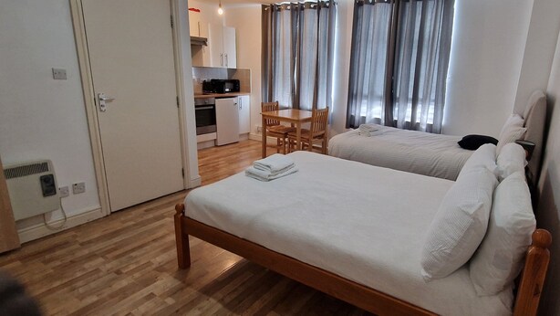 A hotel room with a wooden bed, a small kitchenette, a dining table, and a view of the outside through curtains. - Urban Retreat Studio Camberwell Zone 2 (London)