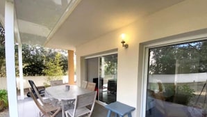 A covered patio area with a round table and chairs, a small stool, and a sliding glass door leading to a garden. - Appartement T3 - 50M DE LA Plage - Rupione (PIETROSELLA)