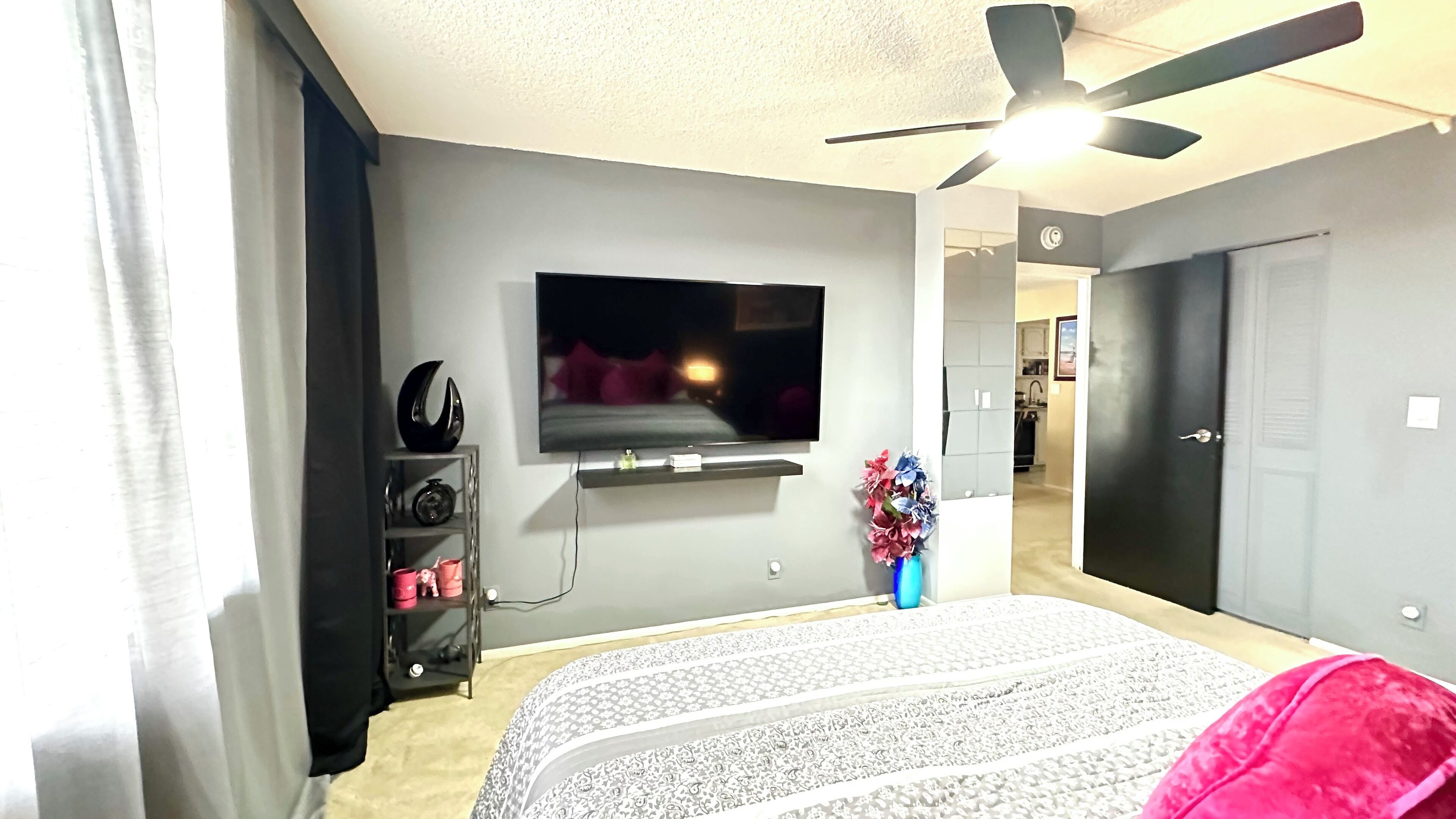 Ein Schlafzimmer mit einem Bett, einem an der Wand befestigten Fernseher, einem Deckenventilator und einer Tür zu einem Badezimmer.