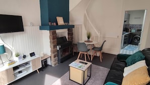 A living room with a fireplace, a TV mounted on the wall, a dining area with chairs, and a kitchen visible through an open door.