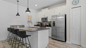 Eine moderne Küche mit weißen Schränken, einem Edelstahl-Kühlschrank, einer Mikrowelle und einer Kaffeemaschine. Drei schwarze Barhocker und Pendelleuchten befinden sich über der Kochinsel.