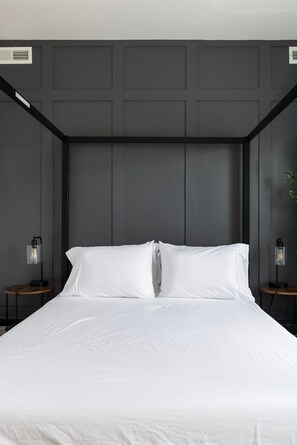 Una cama con ropa de cama blanca, dos mesitas de noche con lámparas y una pared oscura con paneles.