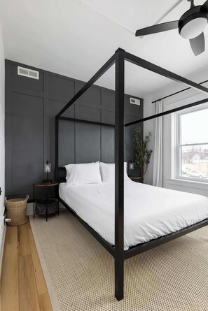 Un dormitorio moderno con cama con dosel, ventilador de techo, mesita de noche, lámpara, ventana con cortinas y vistas al exterior.